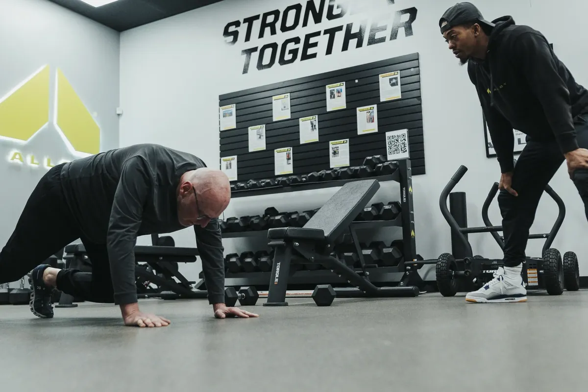 A coach leans in to check an older member's push-up form during a training session